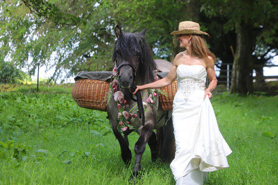 Bride wearing hat walking through Kinclune Estate woodlands with highland pony wearing baskets and flower garland around neck